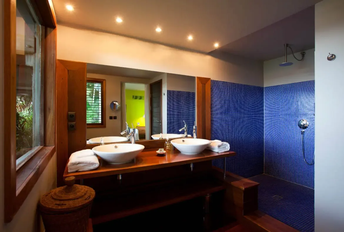 A wooden double vanity with two white vessel sinks sits below a large wood-framed mirror while blue mosaic tiles line the walk-in shower with a rain showerhead. A wooden-framed window reveals lush greenery outside—a bathroom where bold Caribbean blue meets warm tropical wood.