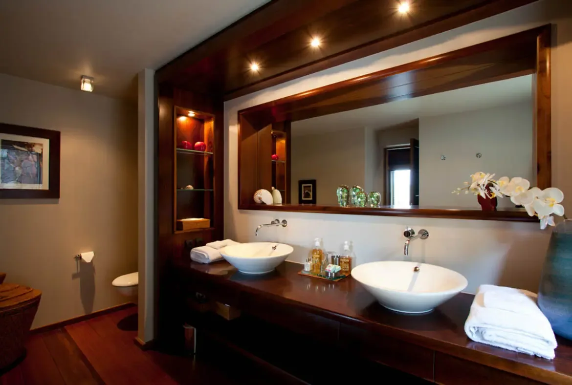 A dark wood double vanity holds two white vessel sinks beside a tray of toiletries and a vase of white orchids. A wide dark-framed mirror reflects warm recessed lighting while glass shelves display red spheres and seashells—a bathroom where tropical elegance meets modern comfort.