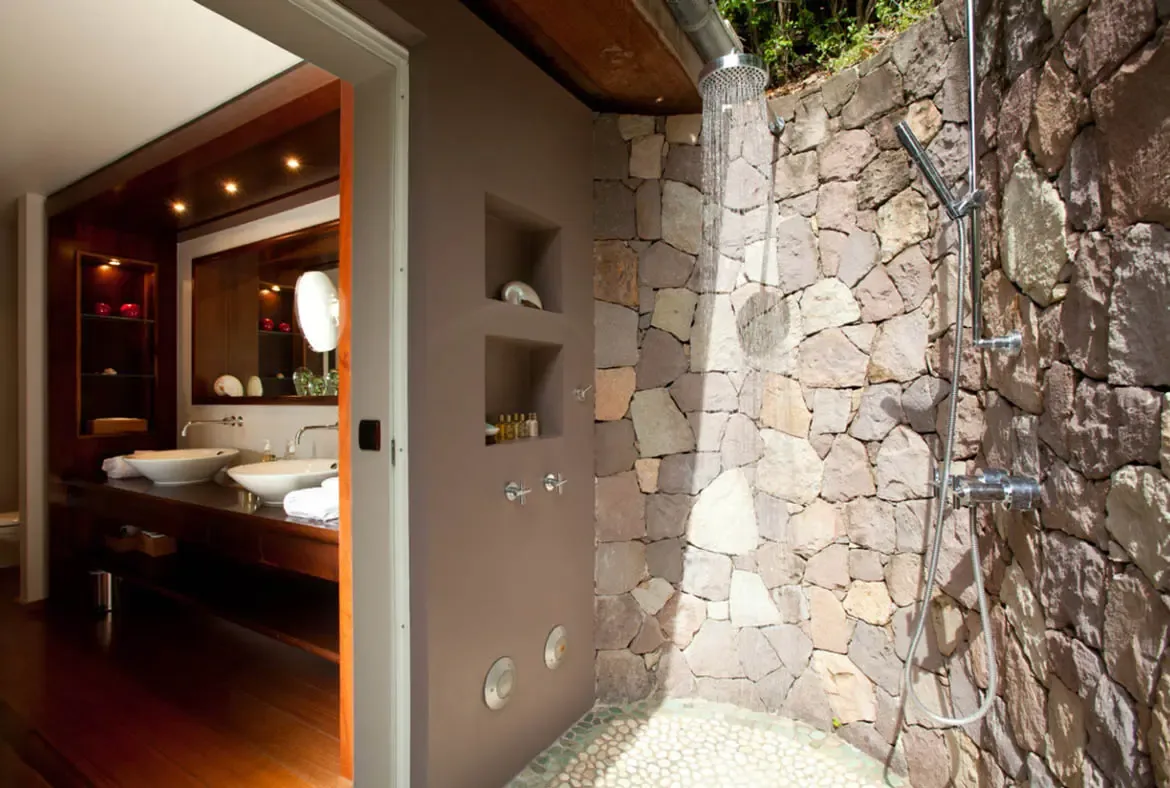 A rain showerhead cascades from the ceiling above a dramatic wall of irregular earth-toned stones with pebble flooring beneath. Through the doorway, a dark wood double vanity with vessel sinks gleams under warm recessed lighting—a spa bathroom that brings the outdoors in.
