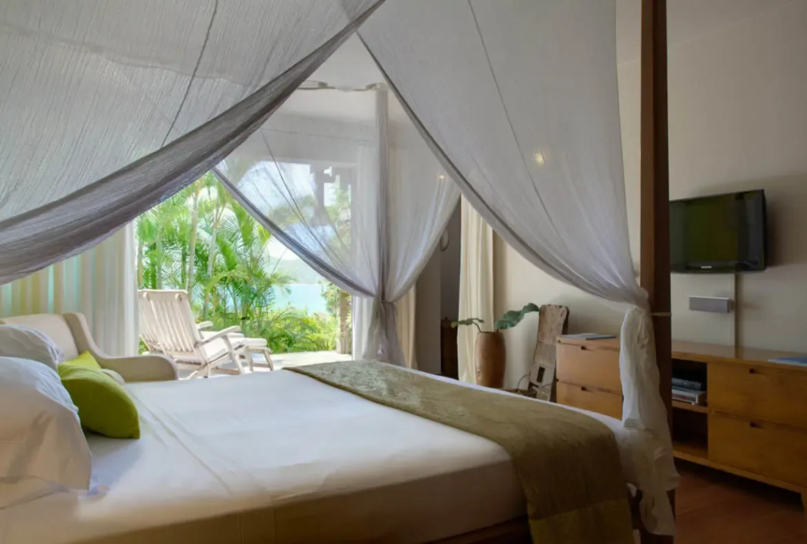 A four-poster bed draped in flowing white canopy netting features a muted green throw and bright accent pillows. Open doors reveal two white lounge chairs on the patio overlooking calm ocean water framed by swaying palms—wake up and step straight into paradise.