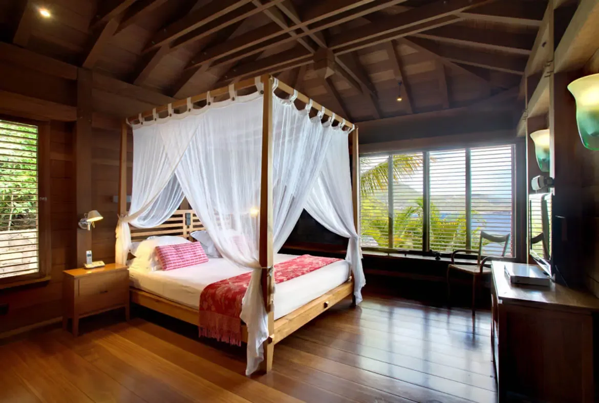 A four-poster bed with a white sheer canopy and a red-patterned throw sits beneath vaulted wooden beams and warm wall sconces. Open louvered windows frame lush palms and a glimpse of ocean beyond—fall asleep to the sound of tropical breezes.