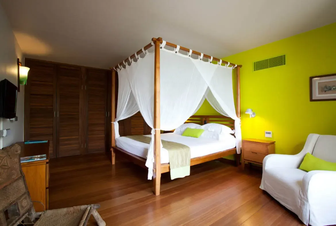 A four-poster bed draped in white canopy fabric sits beside a vibrant lemon-yellow wall with a green accent pillow on a white slipcovered armchair. Dark wooden louvered closet doors and warm wall sconces add character—a bedroom that radiates Caribbean sunshine even indoors.