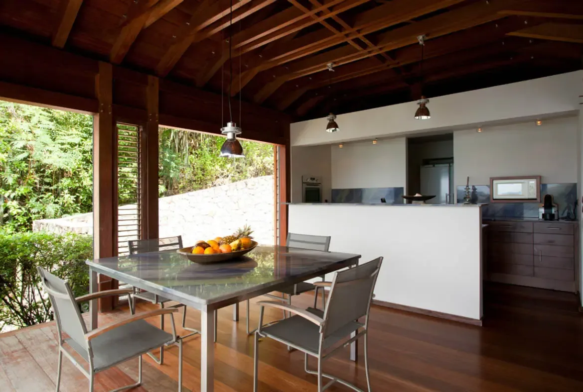 A glass-topped dining table with a vibrant fruit bowl of oranges, lemons, and pineapple sits beneath metallic pendant lights and exposed wooden beams. Open louvered panels connect the dining area to tropical greenery while dark wood cabinetry and a sleek breakfast bar complete the kitchen—fresh island mornings start here.