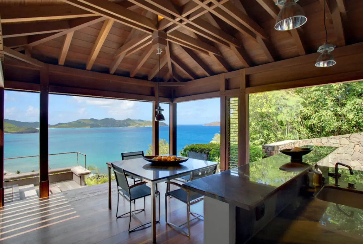 A glass-topped dining table with a bowl of bright oranges sits between a sleek dark kitchen counter and a fully open wall of Caribbean blue. Exposed wooden beams and striped sunlight frame the panorama of turquoise water and green islands—every meal here comes with a horizon-wide view.