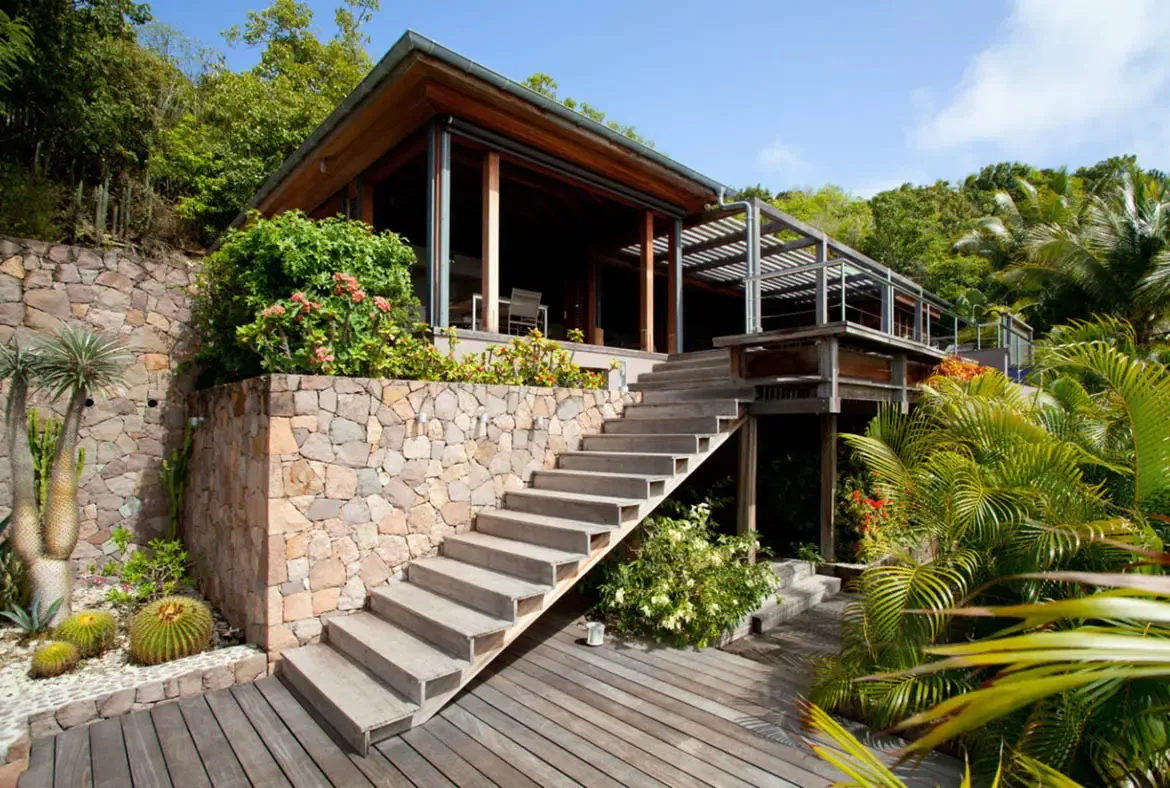 Wide wooden stairs ascend from a lower deck past a stone retaining wall toward the glass-walled main living area. Pink flowering bushes and barrel cacti line the hillside while palms tower above—modern tropical architecture blooming with Caribbean color.
