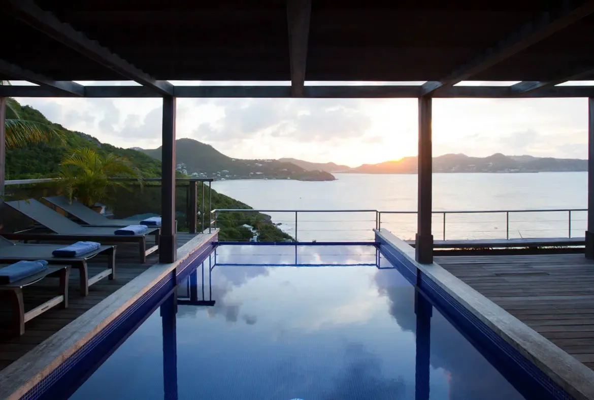 The infinity pool mirrors the golden sky as the sun hovers near the horizon beyond lush green hills and distant islands. Blue-toweled lounge chairs rest under a dark wooden pergola while the glass railing dissolves the boundary between pool and ocean—sunset at Samsara is unforgettable.