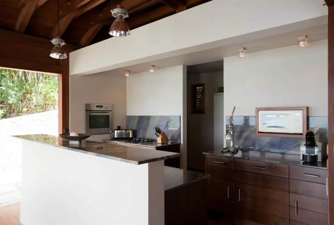 A dark stone island with white cabinetry anchors this open kitchen beneath vaulted dark wooden beams and metallic pendant lights. A marbled blue-gray backsplash complements the walnut cabinets while an espresso machine and stand mixer await—cooking here feels like a Caribbean masterclass.