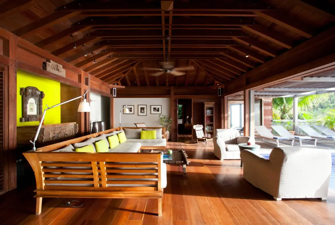 A wooden sofa with light cushions and lime-green pillows sits beneath vaulted dark beams beside a bold yellow accent wall with carved artwork. Open to the tropical breeze and white sunbeds beyond, this airy living room blends vibrant island color with laid-back luxury.