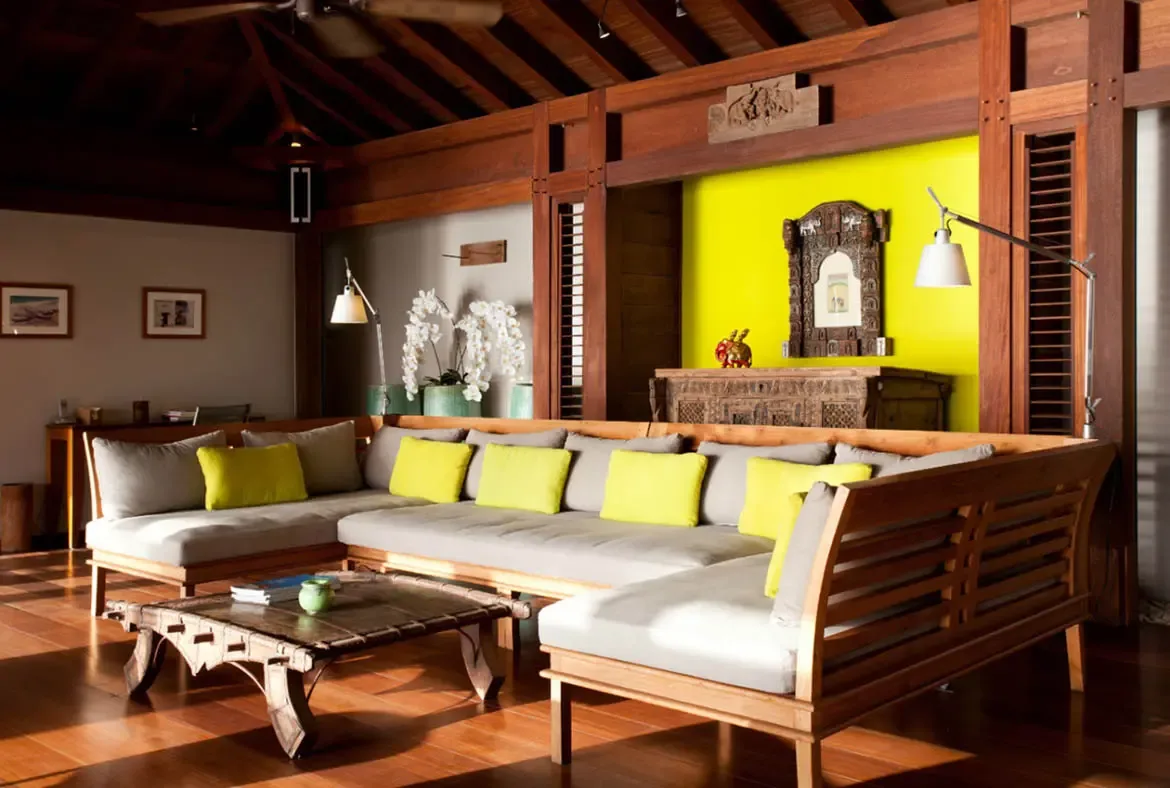 A large wooden-framed sectional with yellow throw pillows anchors this tropical-luxe living room beneath exposed beams and a ceiling fan. A carved wooden cabinet and sculptural wall art pop against the bright yellow accent wall while orchids and polished wood floors soften the scene—warm, stylish, and deeply island-inspired.