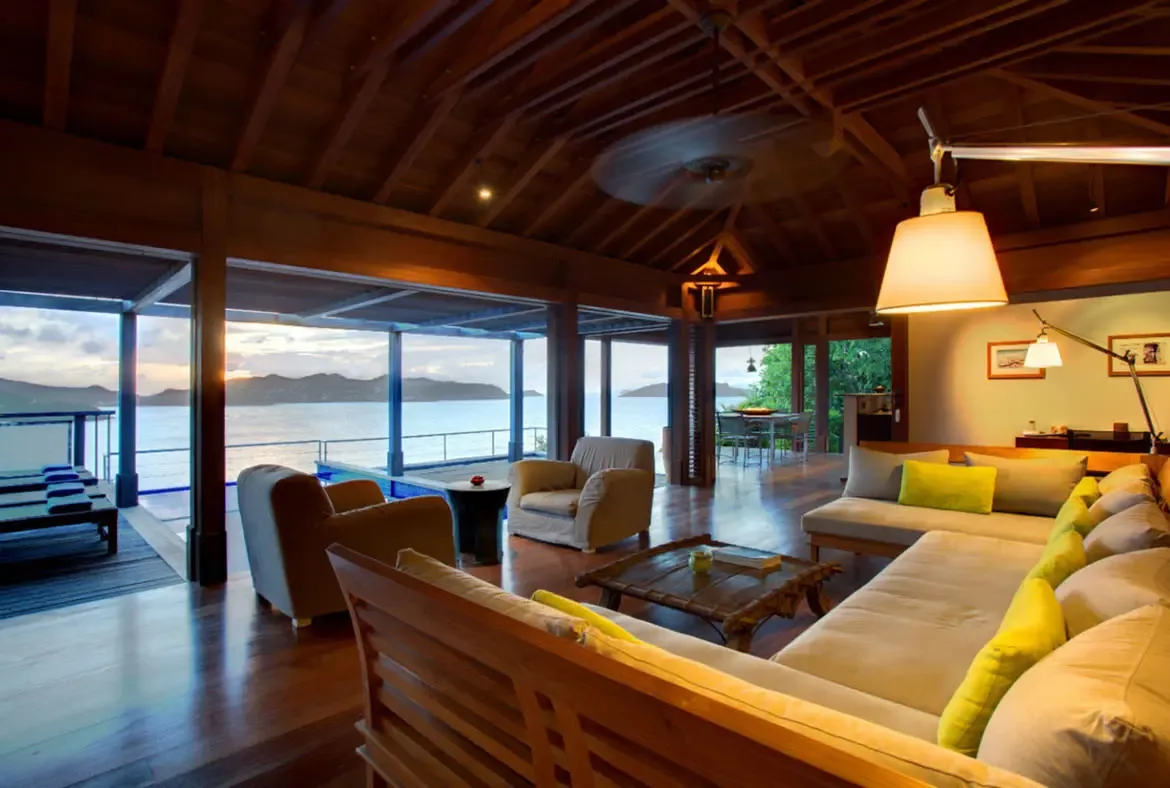 A spacious L-shaped sofa with yellow throw pillows anchors this vaulted living room with exposed wooden beams and polished dark floors. Open views reveal the pool deck, lounge chairs, and ocean glowing at golden hour—a living room made for sunset conversations.