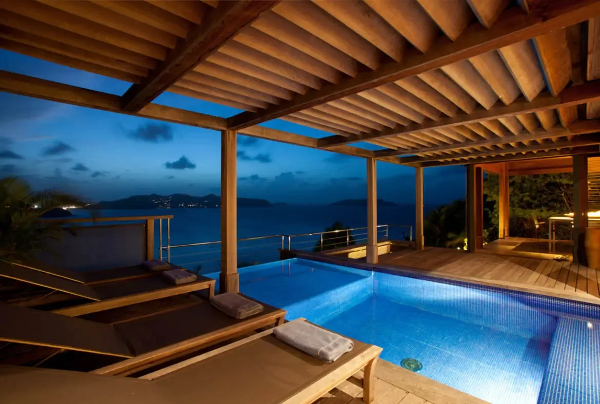 Blue-lit pool water glows beneath a slatted wooden pergola as lounge chairs with rolled towels line the deck. Beyond the railing, the twilight ocean stretches to distant islands while warm interior light spills through open doors—an evening swim with a Caribbean panorama.