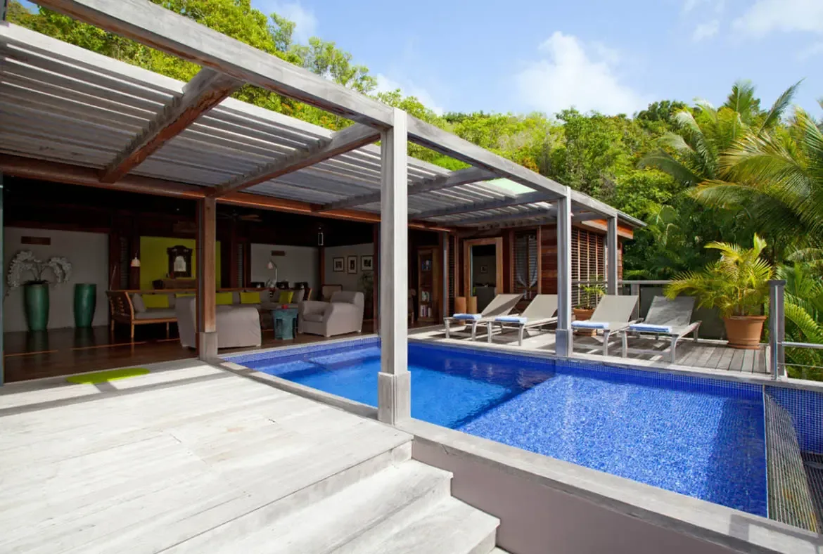 A brilliant blue mosaic pool gleams on a light wooden deck where four cushioned lounge chairs await. A slatted wooden pergola shelters an outdoor living room with sofas and green vases while open doors merge the interior with the tropical garden—seamless indoor-outdoor paradise.