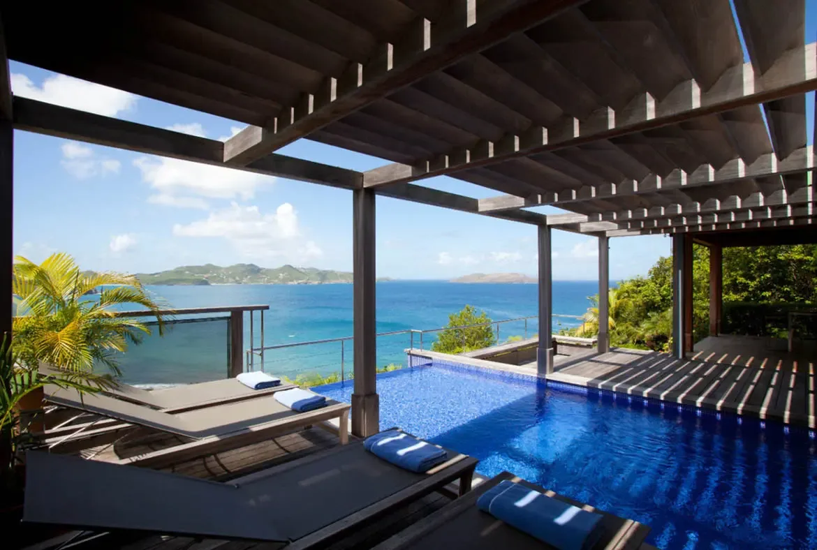 A crystal-blue pool reflects sunlight beneath a slatted wooden pergola where dark lounge chairs await with rolled blue towels. Beyond the pool edge, turquoise ocean stretches to distant islands under a brilliant sky—this is the view that makes Pointe Milou legendary.