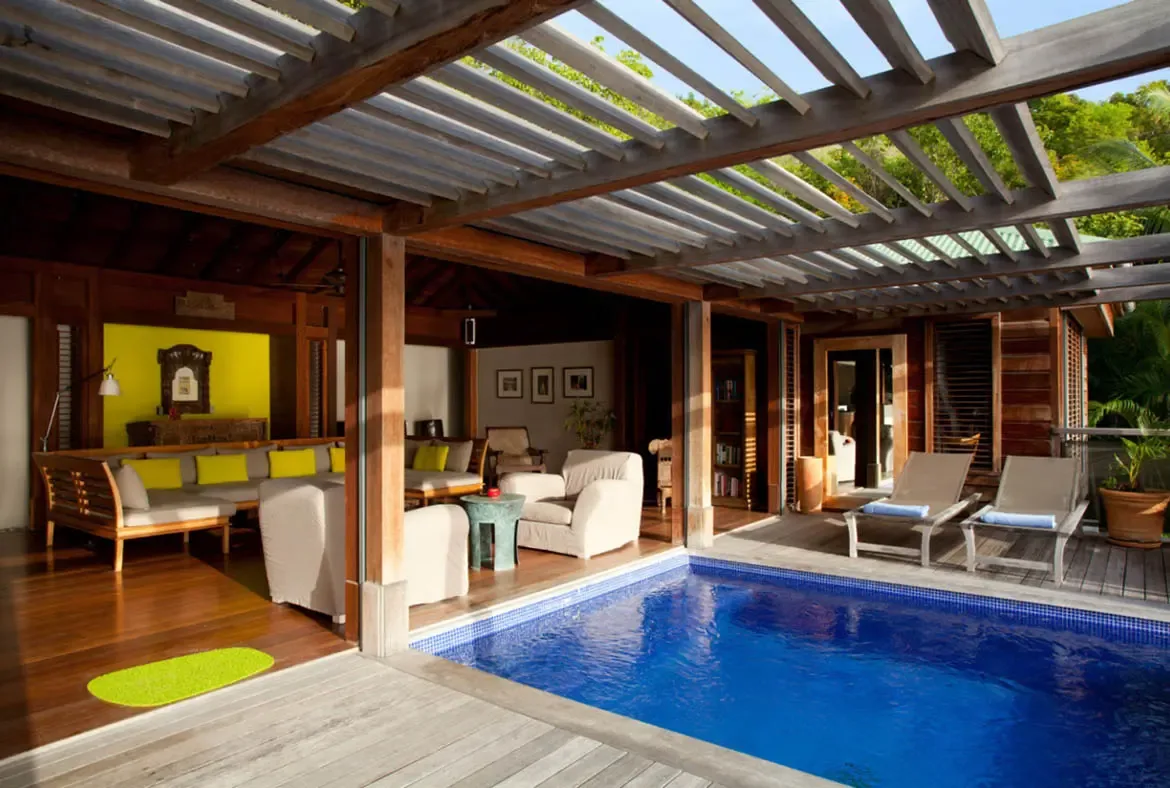 A vibrant blue pool reflects sunlight beside a wooden deck with lounge chairs and blue towels. Under the slatted pergola, a yellow-cushioned sofa and white armchairs create a conversation nook around a turquoise side table while a yellow accent wall with artwork glows beyond—tropical living at its most joyful.