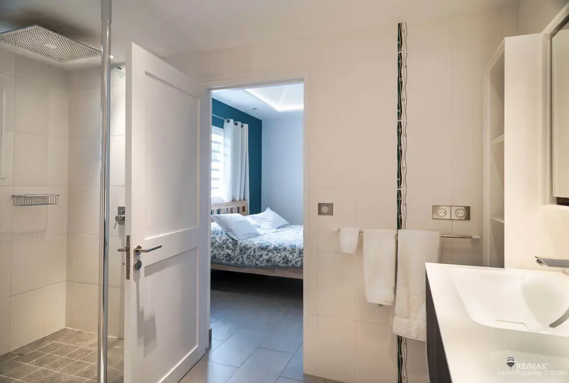 A modern bathroom with grey tile shower and white vanity opens through a door to a bedroom with a blue-patterned bed and a teal accent wall, connecting clean design with coastal comfort.