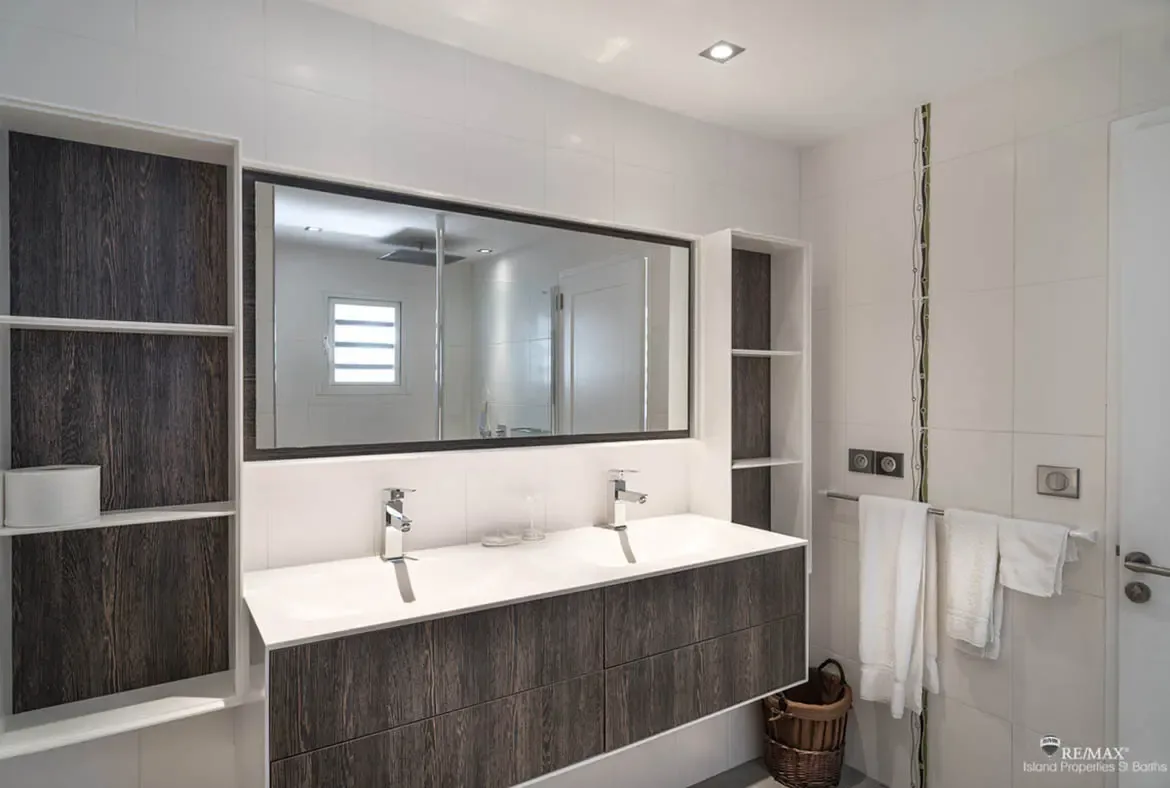 A floating double vanity with dark wood drawers and white integrated sinks sits under a framed mirror, where chrome faucets and built-in shelving create a sleek modern bathroom.