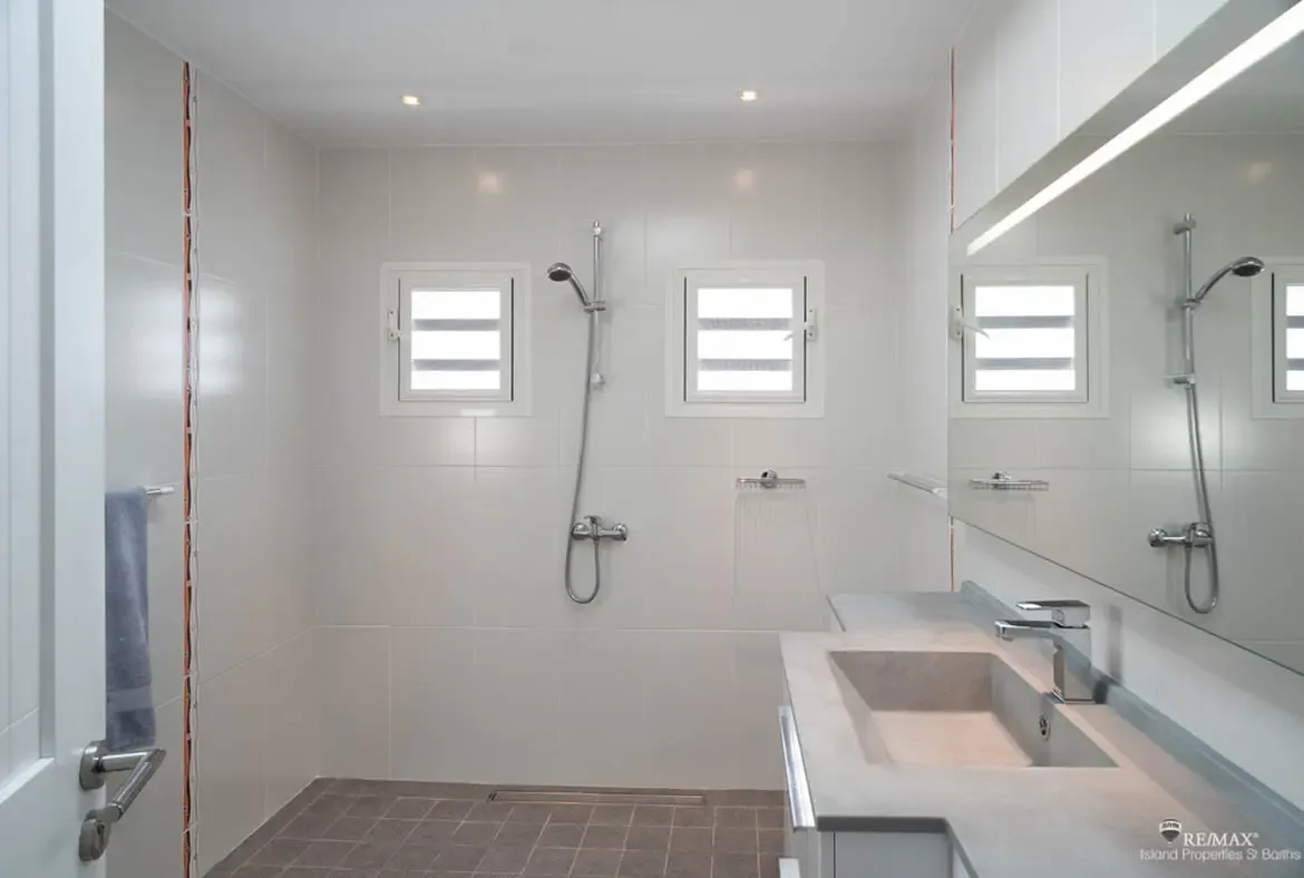 A minimalist bathroom with large grey tiles and a stone vanity features a chrome rainfall shower and louvered windows, where a frameless mirror doubles the sense of space.