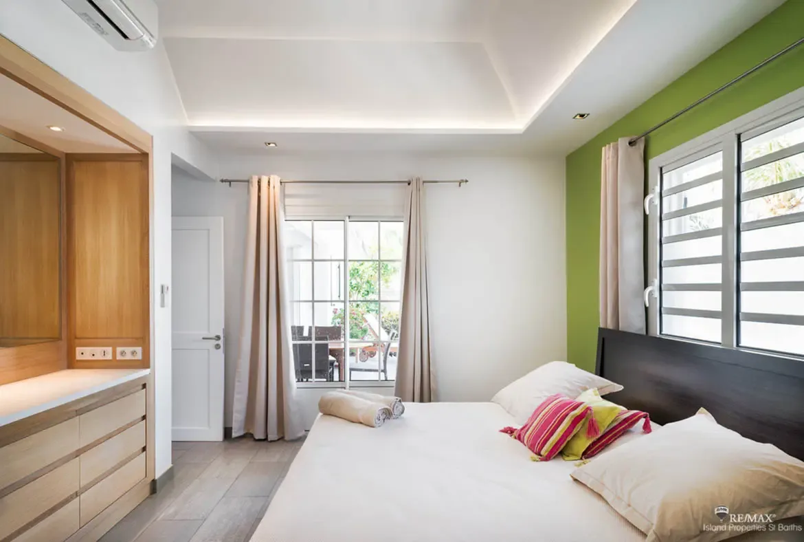 A white bed with a colorful striped pillow sits before a lime-green accent wall, where open doors reveal a private patio with wicker furniture and tropical greenery.