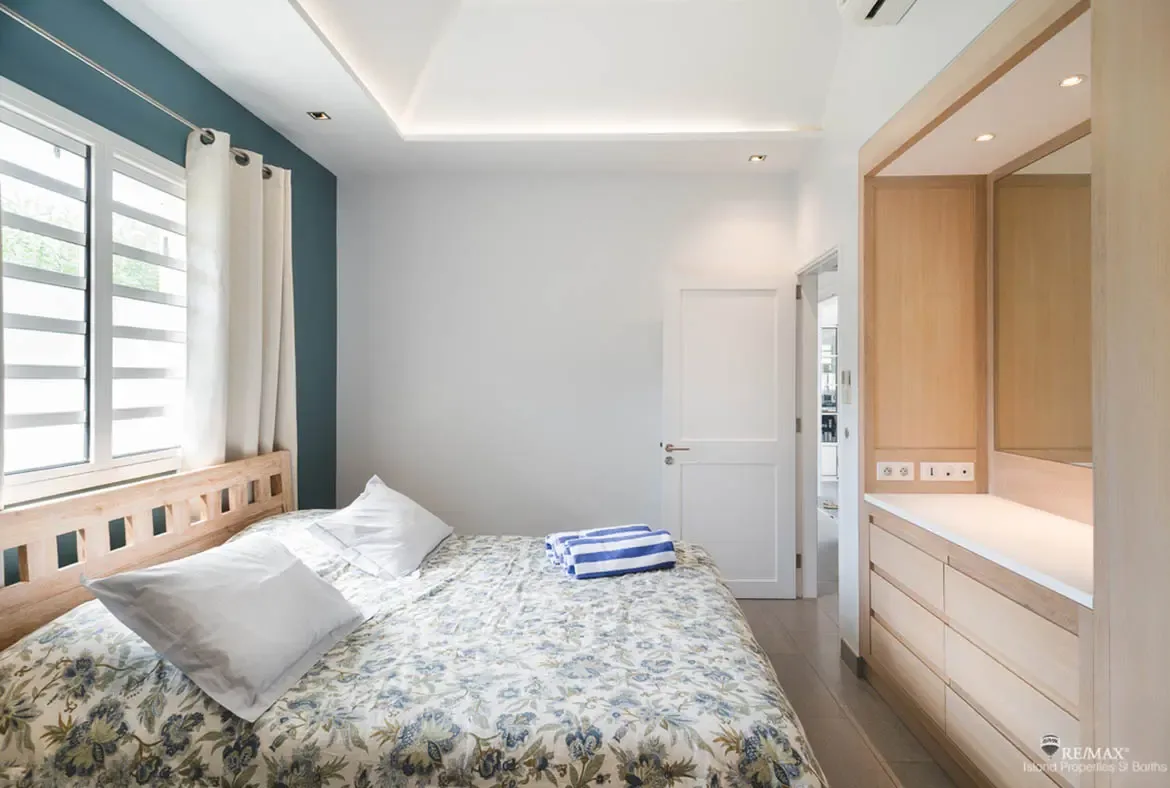 A light wood bed with yellow floral bedding sits before a deep teal accent wall, where a built-in wardrobe with mirror and recessed lighting adds modern elegance.