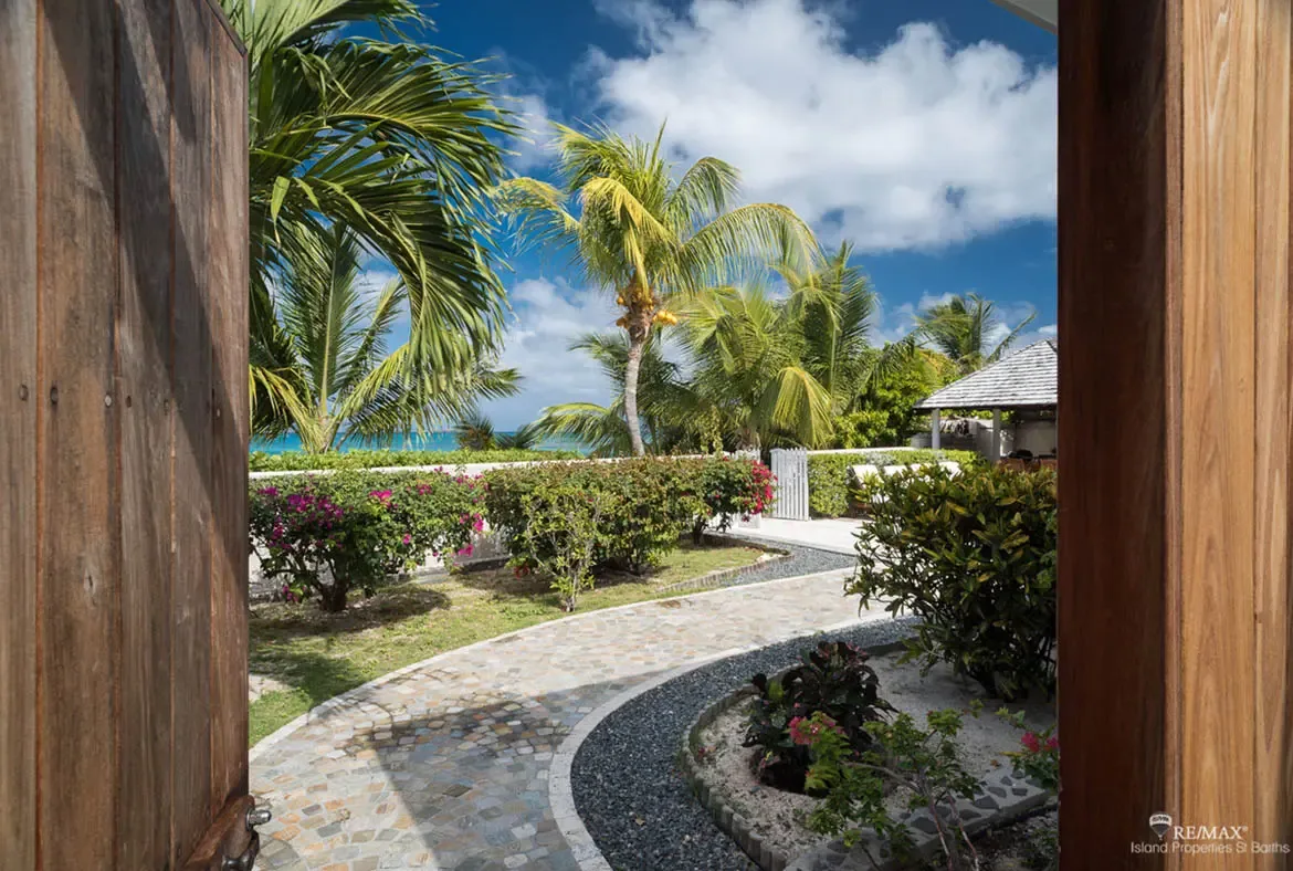 Dark wood doors frame a curved stone path bordered by river pebbles, where pink bougainvillea and coconut palms lead to a white picket fence and turquoise ocean beyond.