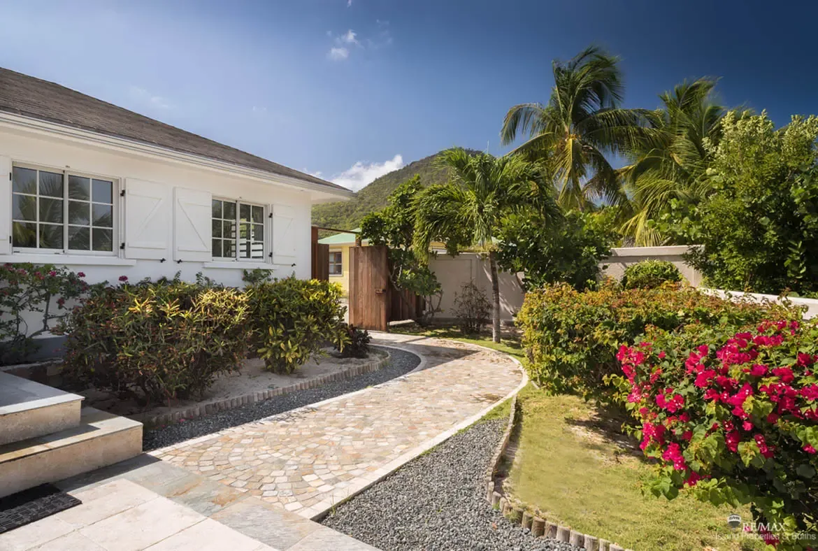 A curved cobblestone path leads through pink bougainvillea and green hedges to a white shingle-roofed villa, where coconut palms rise against a green hillside under a blue sky.