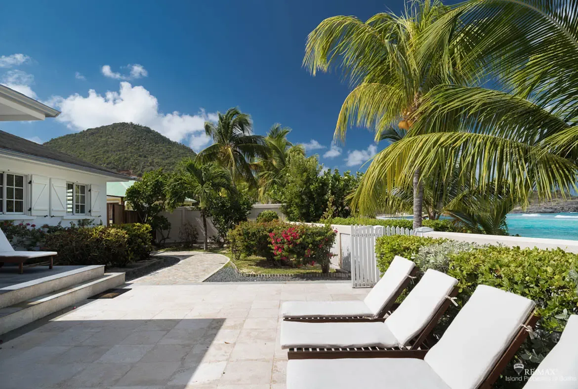 White loungers sit on a sun-drenched stone patio beside steps and a curved path, where pink bougainvillea, coconut palms, and a white gate lead toward turquoise ocean and a lush green mountain.