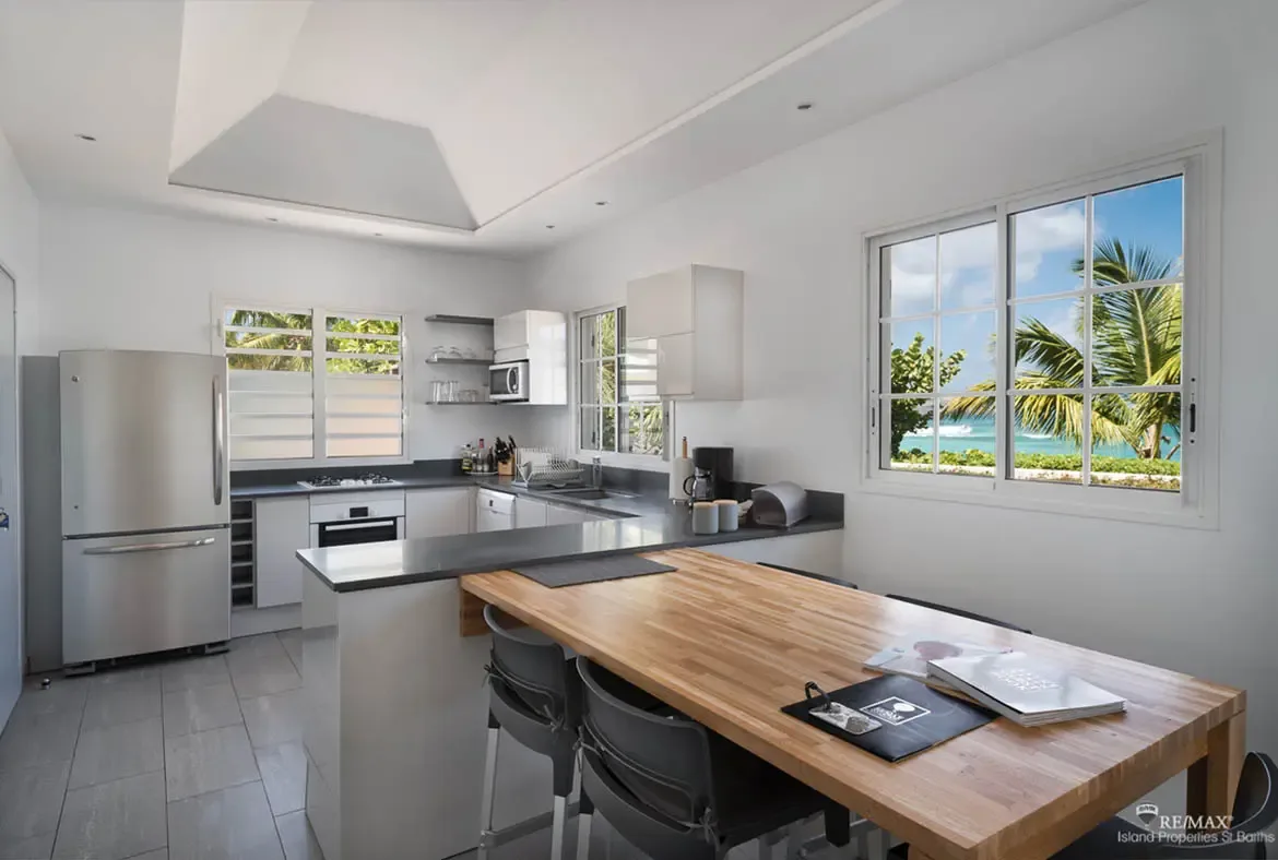 White handle-less cabinets and dark charcoal countertops form an L-shaped kitchen, where two black stools sit at a wood breakfast bar and large windows frame turquoise ocean and palm trees.