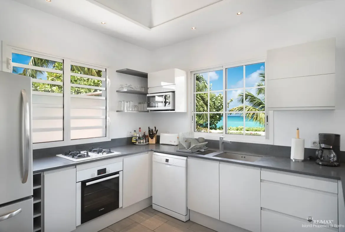 A sleek white kitchen with a gas stovetop and wine rack faces louvered windows, where turquoise ocean and swaying palms fill the frame beyond dark countertops and minimalist shelving.