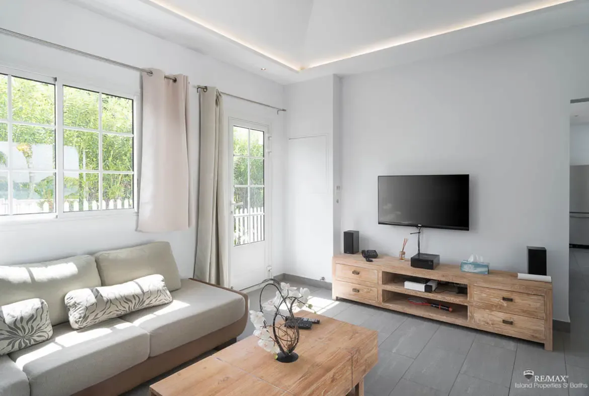 A light taupe sofa with botanical pillows faces a wooden media console under a tray ceiling, where white orchids on a coffee table add elegance to a bright coastal living room.