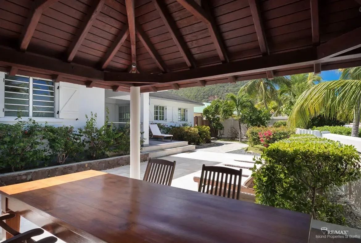 A dark wood dining table with matching chairs sits under a vaulted pavilion with exposed beams, where white pillars frame a lush garden path and the white villa beyond.