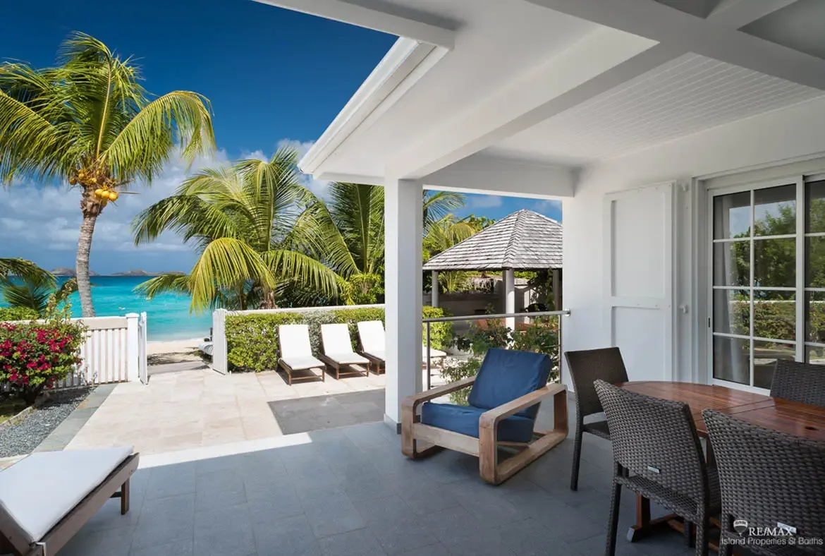 White loungers line a stone terrace beside pink bougainvillea and a picket fence, where palm trees frame turquoise ocean and a distant green hillside.