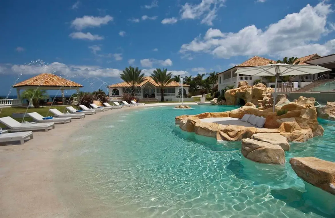 An aerial view shows a turquoise lagoon pool with a beach entry and rock island with a white umbrella, where white loungers and a terracotta gazebo line the green lawn.