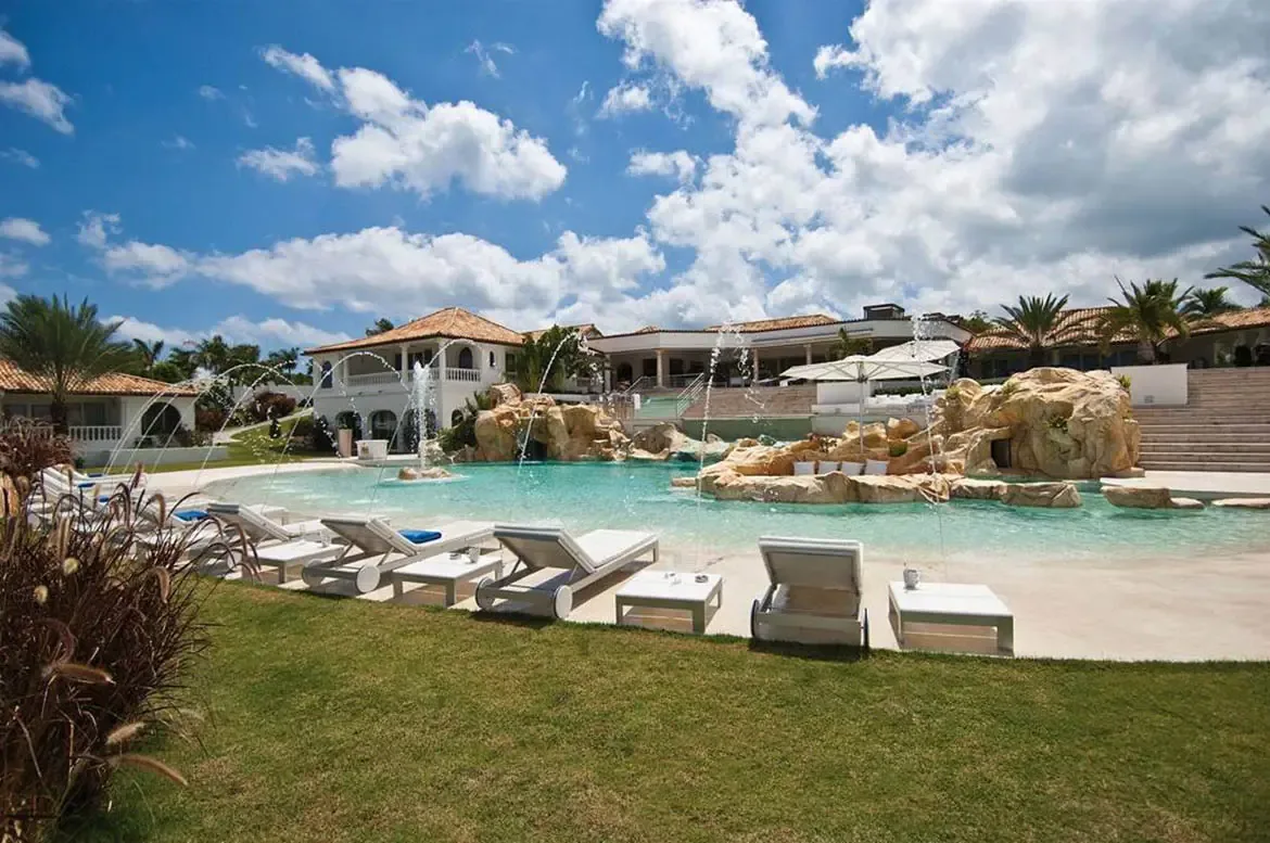 An aerial view of a sprawling white villa with terracotta roofs beside a lagoon-style pool with rock islands and beach entry, where white loungers line a green lawn above the ocean.