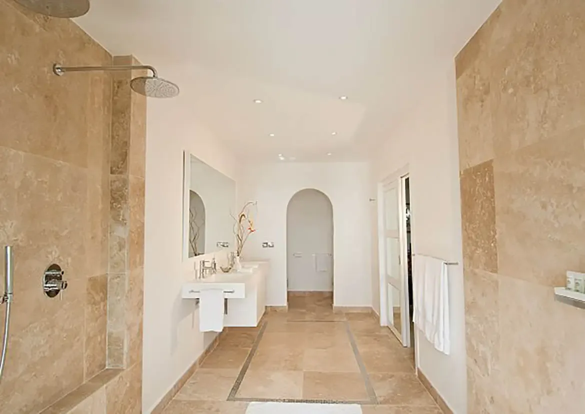 Beige stone tiles cover a walk-in rain shower and a white floating vanity with chrome faucets, where dried reeds in a vase and a frameless mirror fill the bright space.