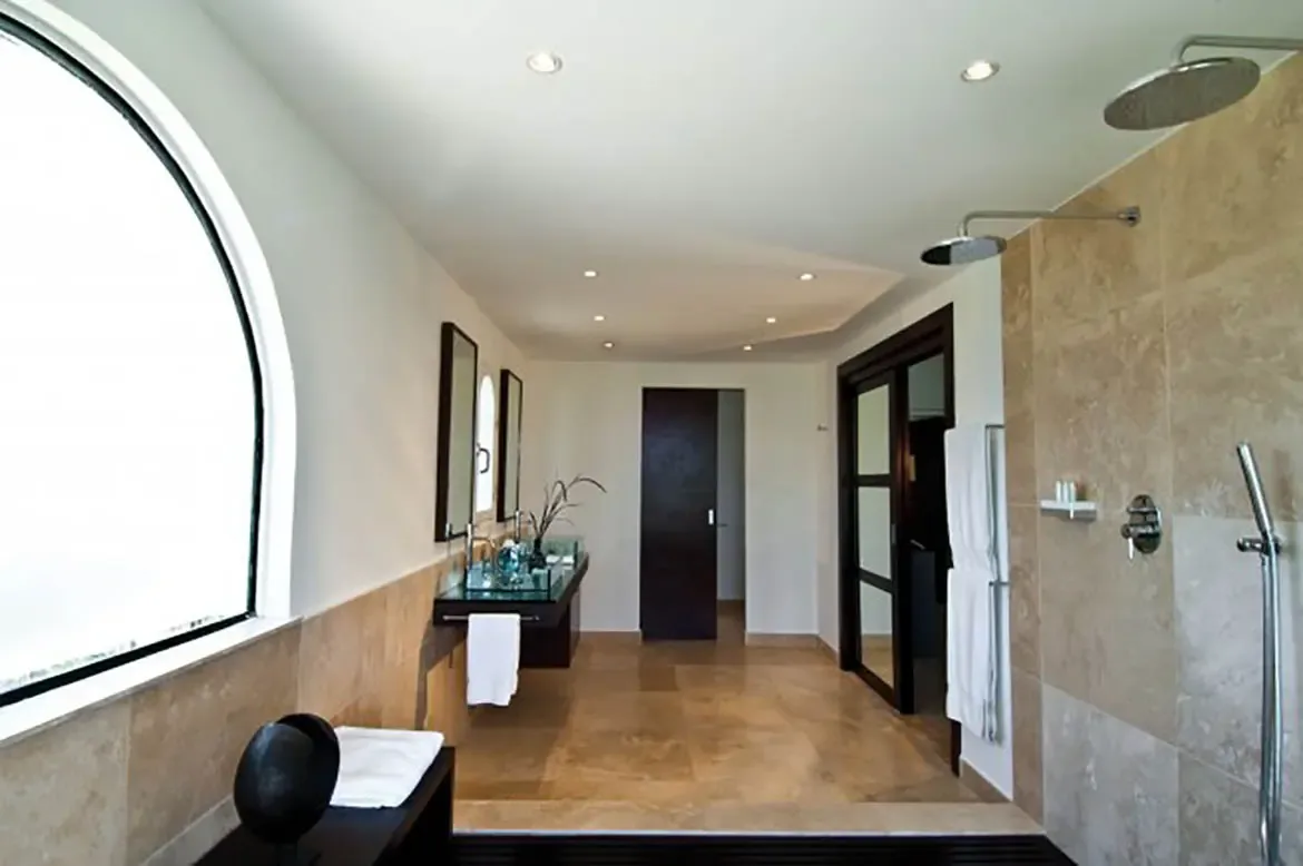 Two light-blue glass vessel sinks sit on a dark wood vanity beneath tall mirrors, where dual rain showerheads and an arched window with bright light fill the stone-tiled bathroom.