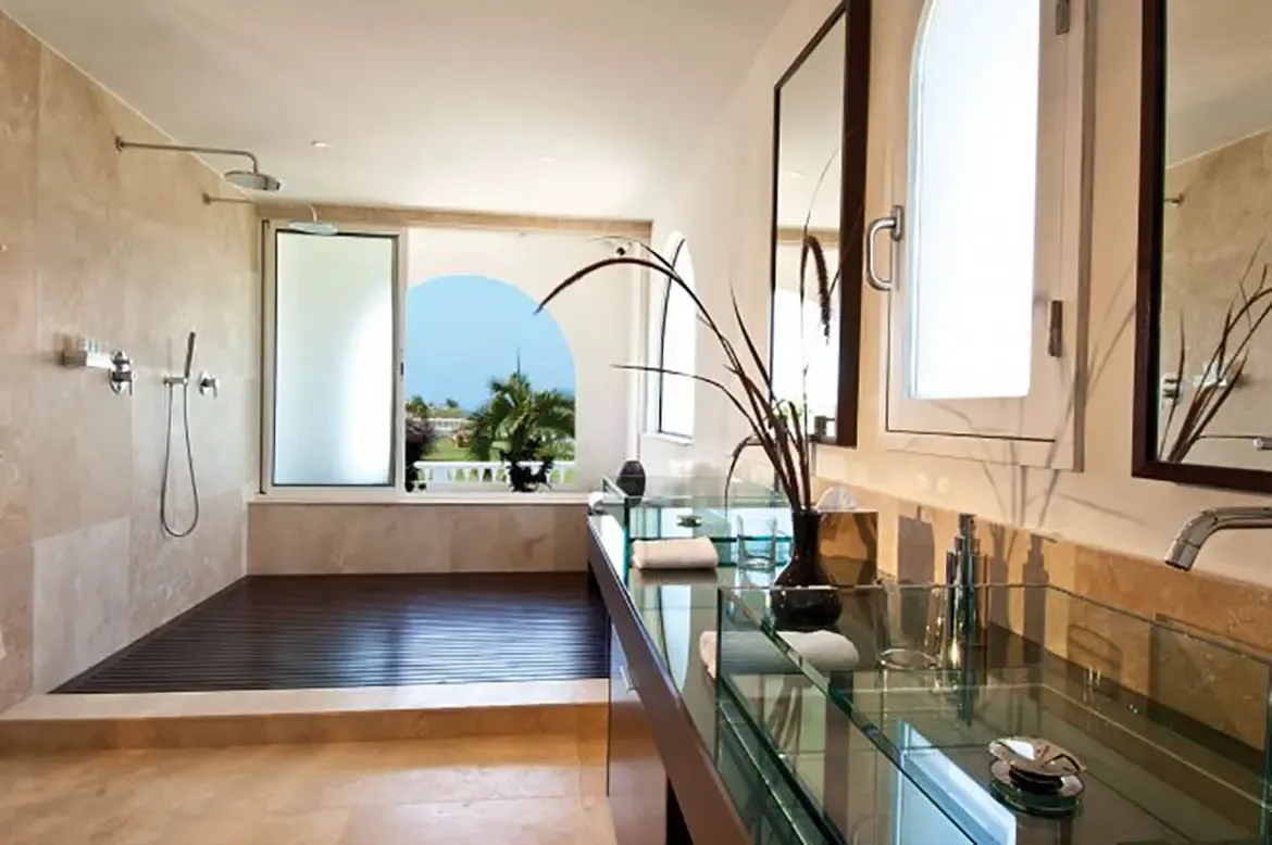 Two glass vessel sinks on a thick glass counter sit between dark-framed mirrors, where a double shower with wood slats and an arched window reveal palms and ocean.