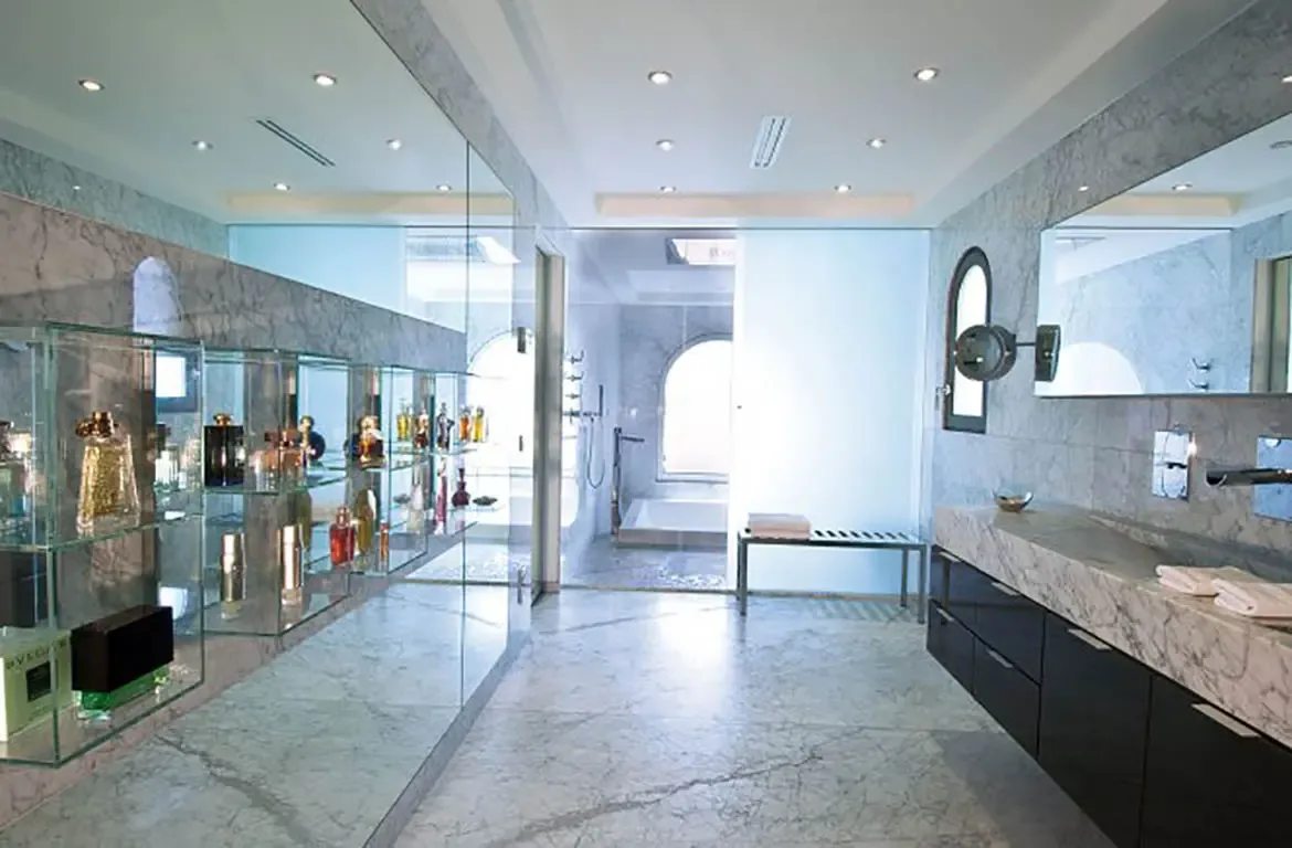 Grey-veined marble covers a floating vanity with chrome fixtures beside a perfume alcove, where a soaking tub sits under an arched window beyond a glass partition.