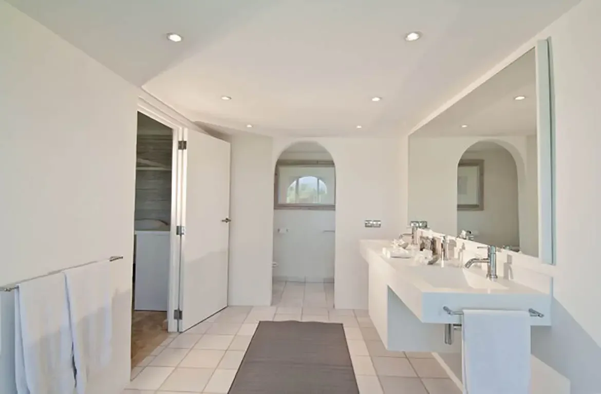 A long white floating vanity with integrated sinks and chrome faucets sits beneath a frameless mirror, where an arched doorway leads to a closet and recessed spotlights illuminate the space.