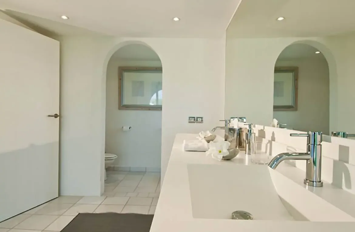 A sleek white double vanity with chrome faucets and a white plumeria flower sits beneath a frameless mirror, where an arched doorway leads to a separate toilet alcove.