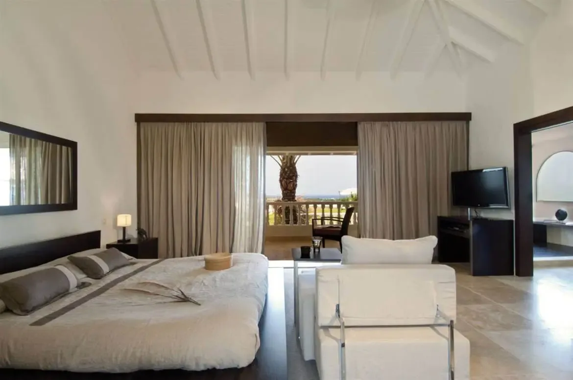 A dark-wood bed with taupe pillows sits under a vaulted ceiling near a chrome chair, where glass doors open to a stone balustrade balcony with palm trees and ocean view.