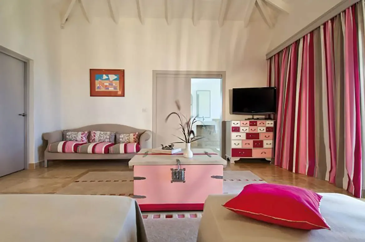 A pink vintage trunk sits between twin beds with grey linens and fuchsia pillows, where a curved grey sofa faces a pink-and-red dresser beneath bold striped curtains.