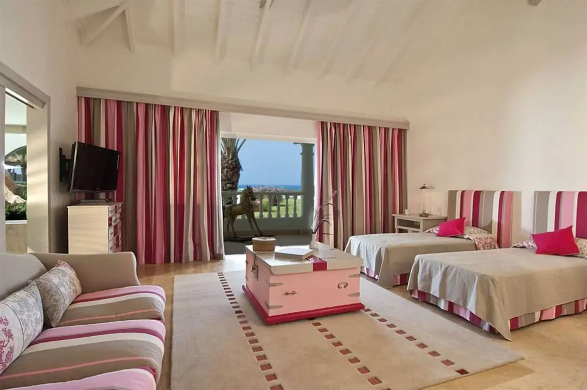 Two twin beds with fuchsia pillows and pink-striped headboards sit under a vaulted ceiling, where floor-to-ceiling pink curtains frame a balcony with a wooden horse sculpture and ocean view.