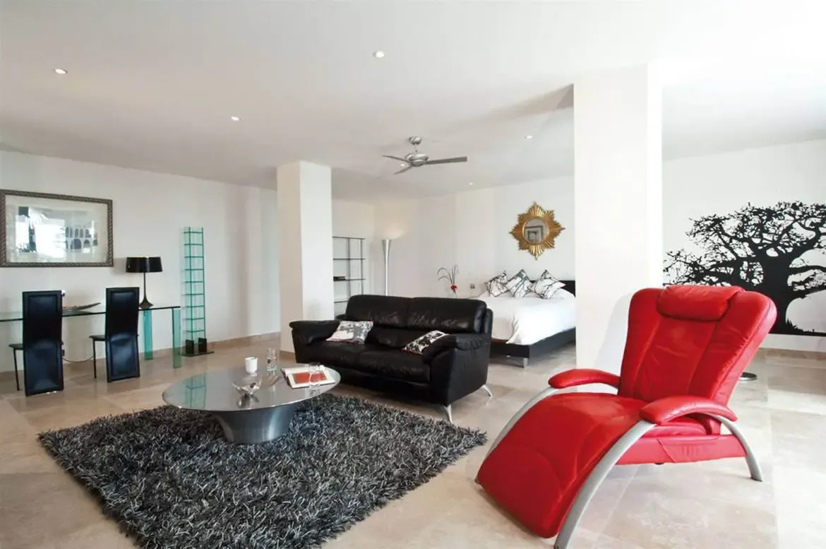 A black leather sofa and red leather chair sit on a dark rug before a white alcove bed with a gold sunburst mirror, where a tree mural and glass desk fill the open-plan suite.