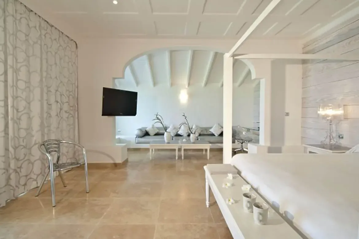 A white four-poster bed with white linens and a wooden bench sits under a white coffered ceiling, where a lattice-pattern curtain and a chrome chair frame a sitting alcove with branch vases.