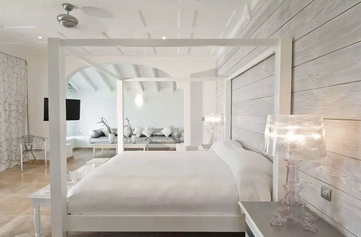 A white four-poster bed with white linens sits against a grey wood-plank wall, where a white arch opens to a sitting area with a grey sofa and branch vases.