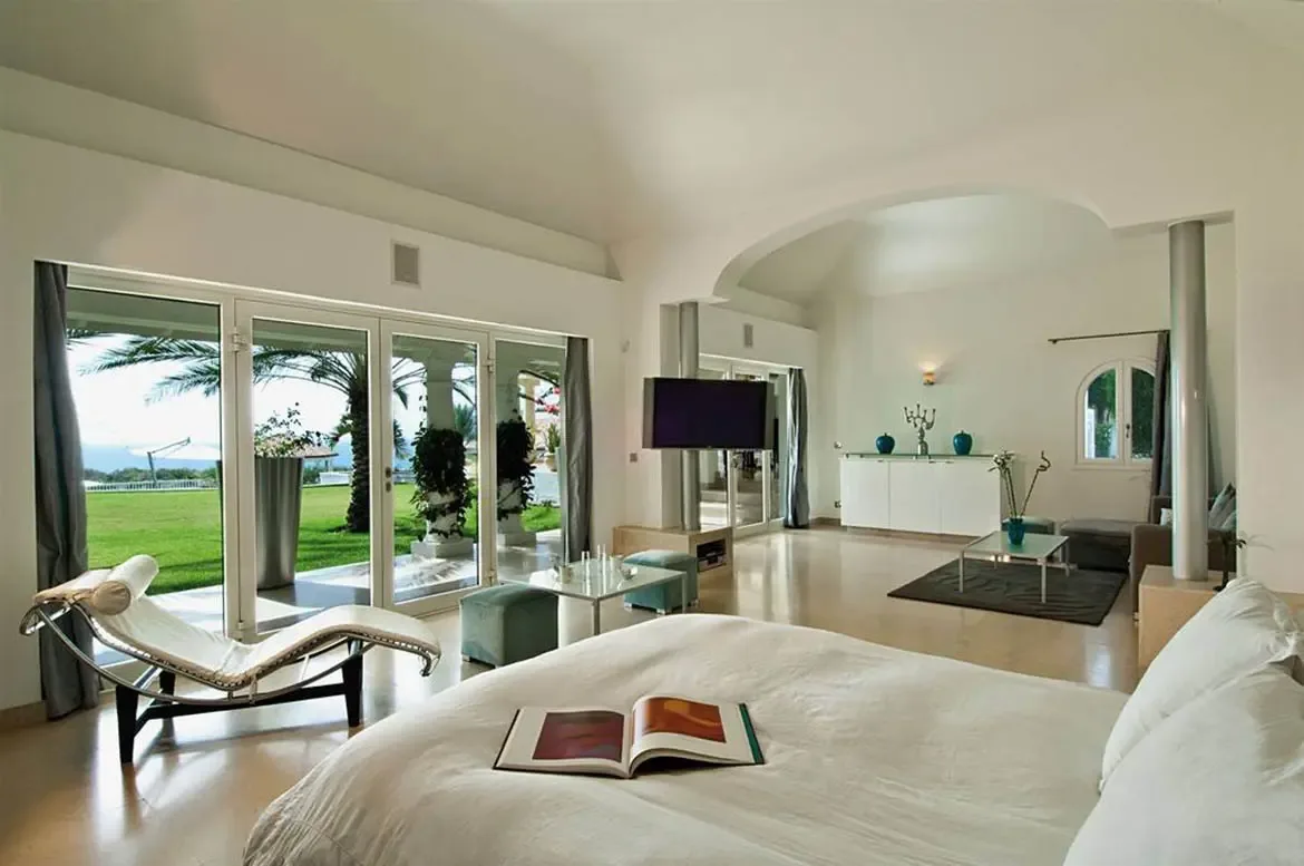 White linens drape a king bed beside a classic white leather chaise lounge, where teal ottomans and a glass table sit before glass doors opening to palms and lawn.