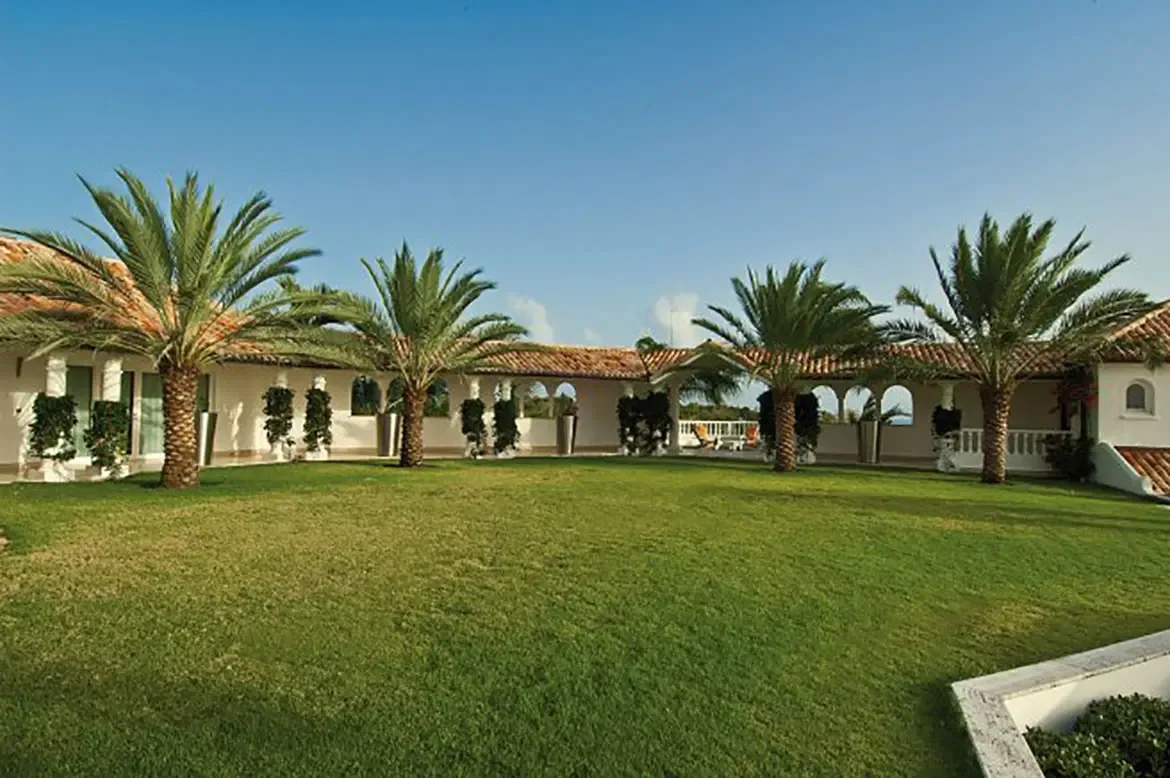 Four palm trees stand on a green lawn framed by white arched walkways and terracotta roofs, where climbing plants accent the Mediterranean villa's courtyard.