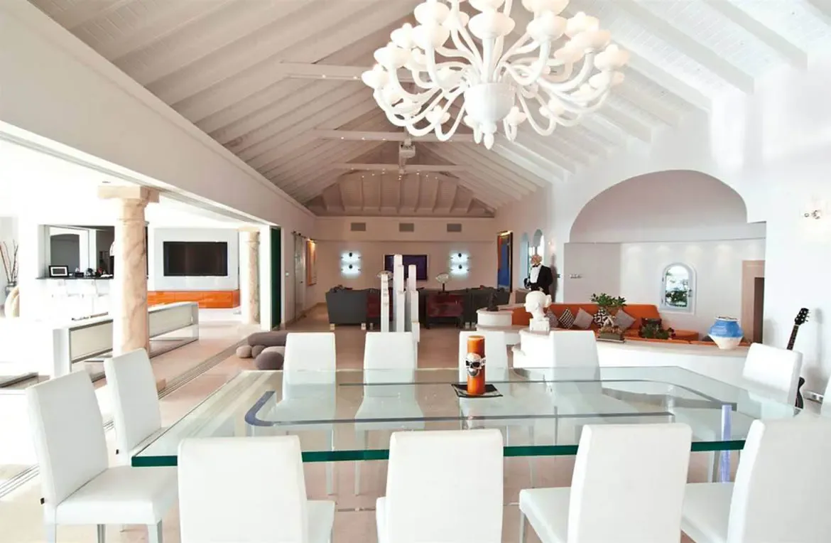 A glass dining table with white chairs and a branching white chandelier sits under a vaulted ceiling, where an orange skull candle and a tuxedo-clad mannequin add avant-garde flair.