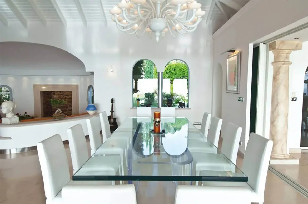 A glass dining table with ten white chairs sits under a branching white chandelier in a vaulted white room, where arched windows reveal lush greenery beyond classical columns.