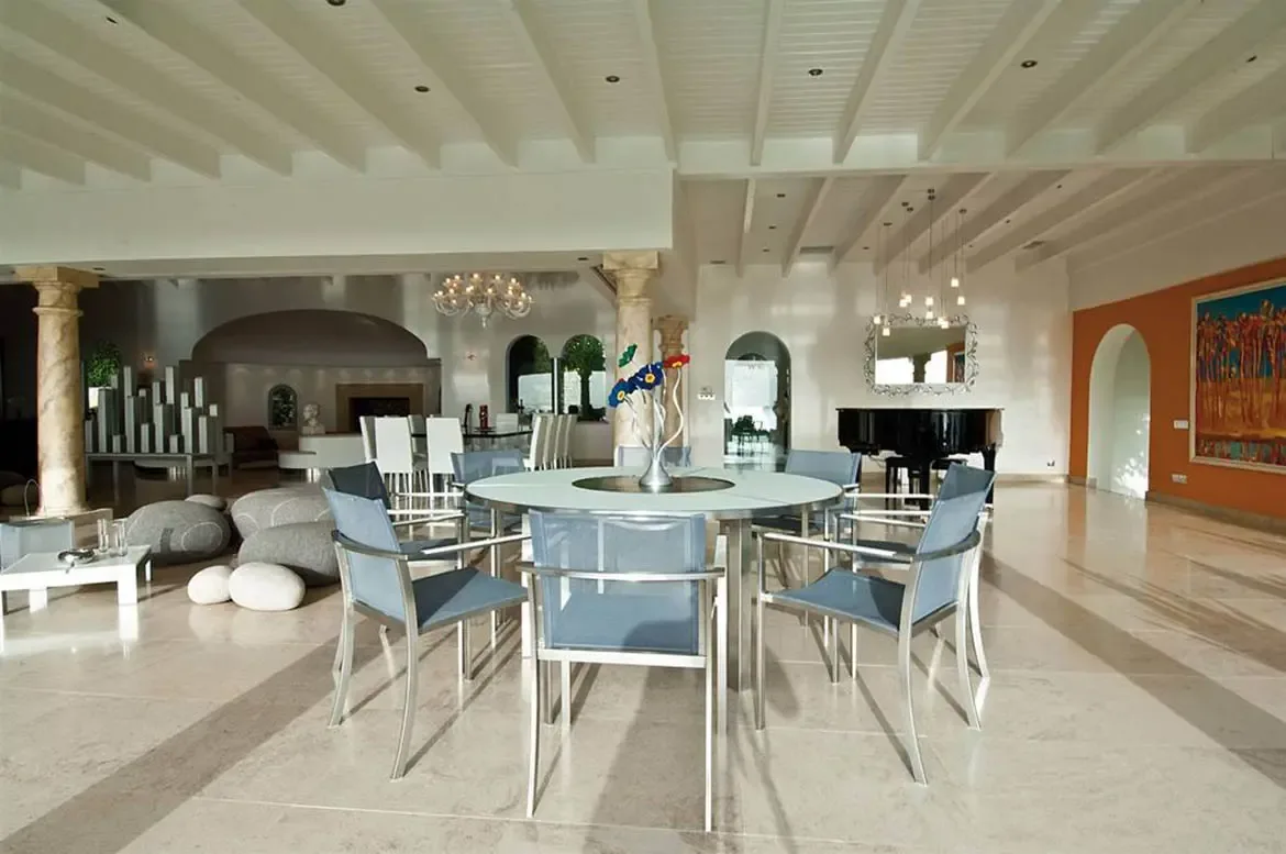 Blue chairs circle a frosted glass table with a blue-and-yellow flower centerpiece, where a black grand piano sits beneath pendant lights near an orange accent wall.